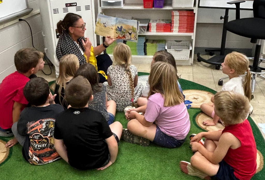 New principal Ms. McClain reads to a group of 10 elementary students seated on the green carpet around her