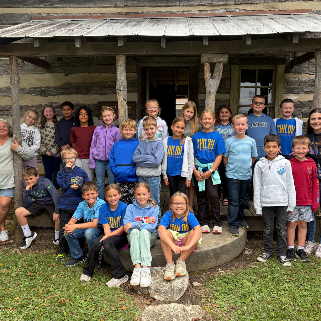 Group photo of 4th graders posing outside of a log cabin on their field trip