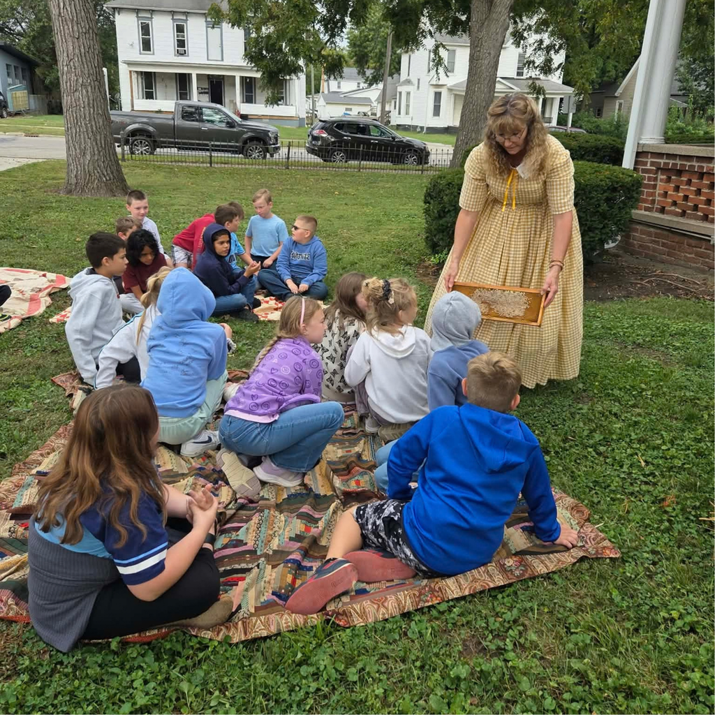 4th grade students sit on the ground outside and listen to a woman in historical garments talk about what life was like in the past