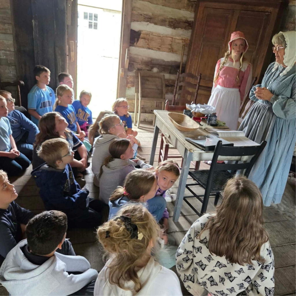 4th grade students sit on the ground in a log cabin and listen to two women in historical garments talk about what life was like in the past
