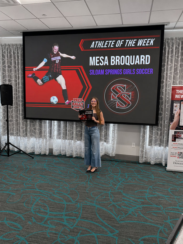 A woman stands in front of a large screen showing a soccer player with "Athlete of the Week" text. She holds a trophy.