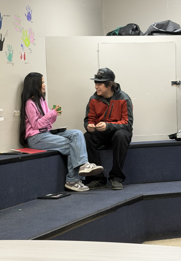 Two individuals sit on a blue platform, engaged in conversation. The person on the left wears a pink shirt and jeans, the other in a black and red jacket.