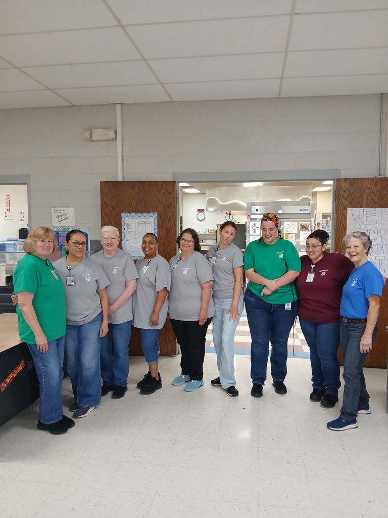 Nine people wearing similar T-shirts and jeans, smiling, and posing for a group photo in a hallway.