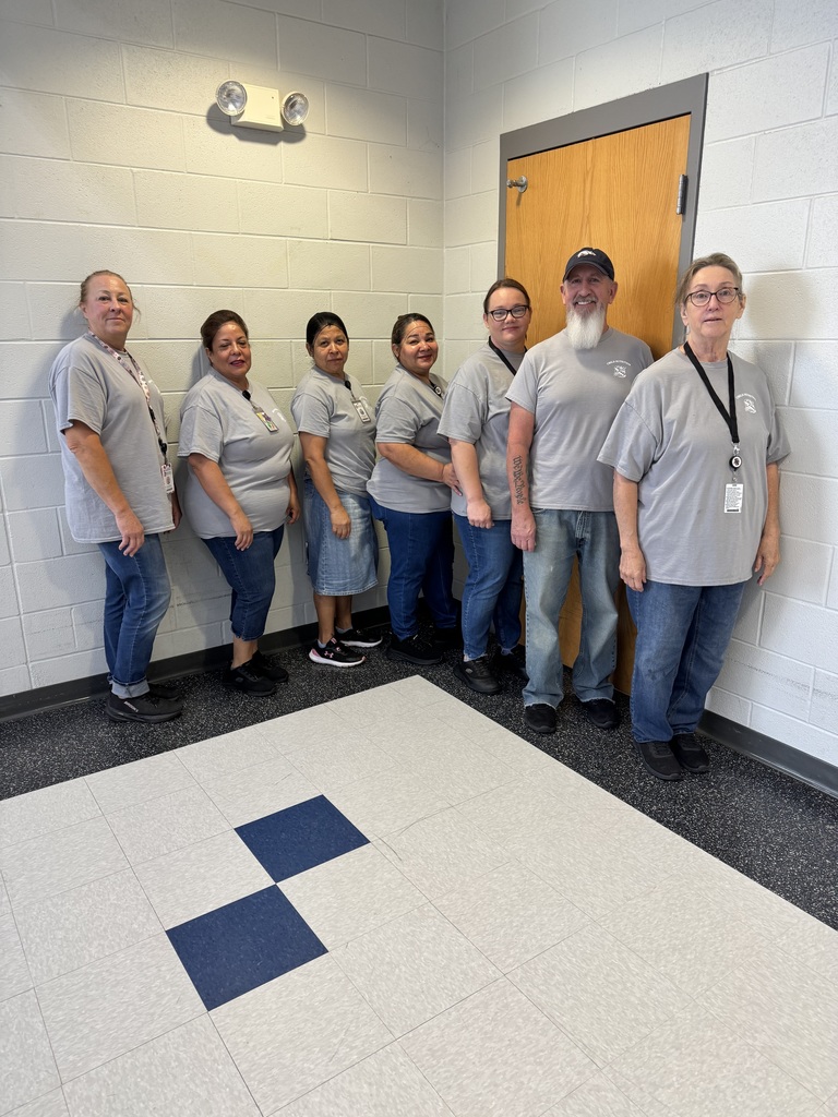 Six individuals in gray uniforms and lanyards pose for a photo in a room with white walls and a blue-patterned floor.