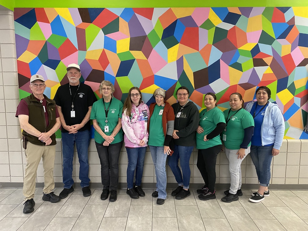 A group of eight people stand in front of a colorful geometric mural. They wear green shirts.