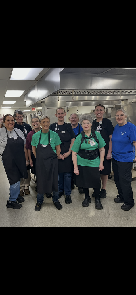 A group of individuals, some wearing aprons and eyeglasses, stands in a row in a kitchen.