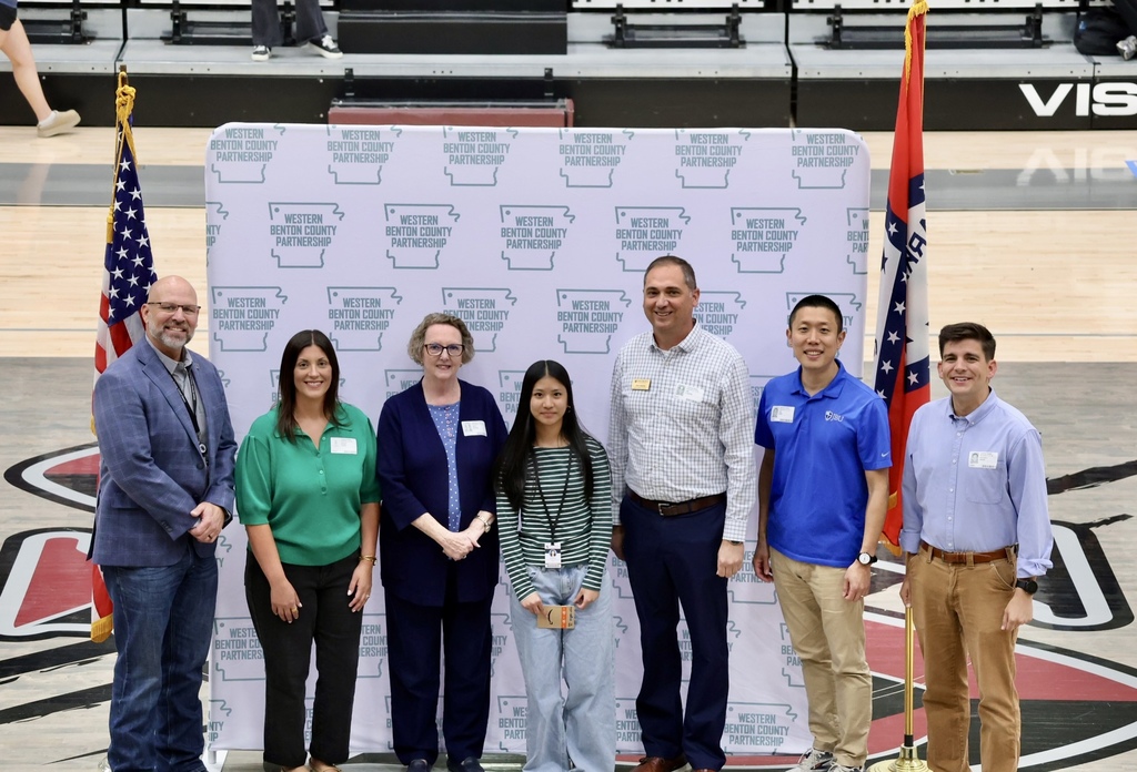 Seven people stand in front of a banner with logos and flags behind them on a basketball court.