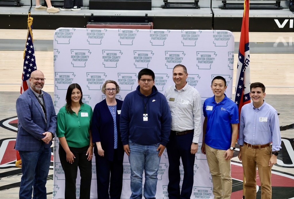 Seven people stand by a sign with logos and flags in the background. They wear varied colors and styles of clothing.