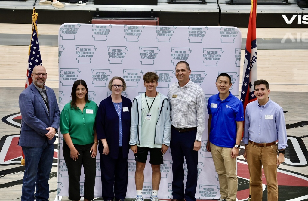 Six adults and a young man stand behind a banner with American flags on a court.