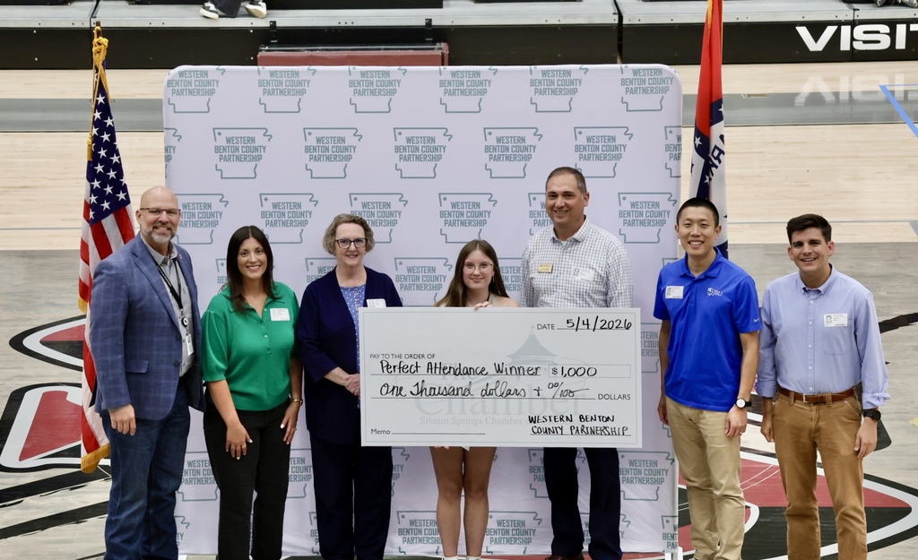 Group of people, some wearing glasses, standing behind a large check. They are on a basketball court with a large wall behind them.