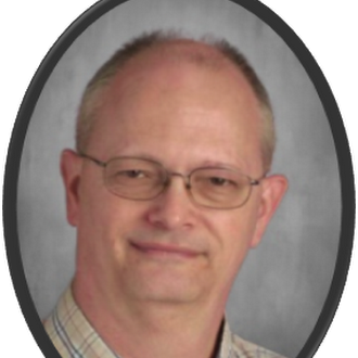 A man with short, light hair and glasses smiles at the camera. He wears a checkered shirt.