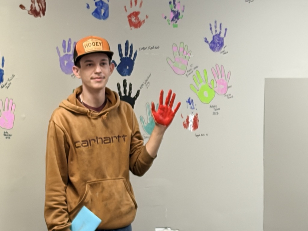 Person in brown hoodie with orange hat and red-dyed hand stands in front of a wall with multicolored handprints.