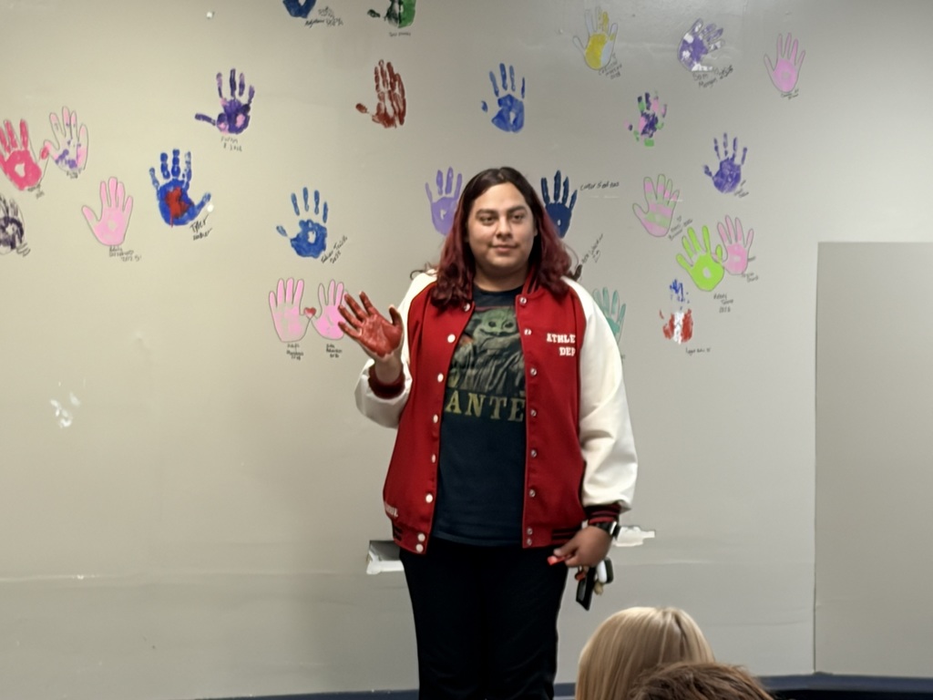 A woman with red hair stands in front of a wall with colorful handprints, wearing a red jacket.