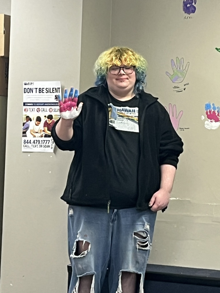 Person with blue hair and glasses stands next to a wall with a poster and handprints.