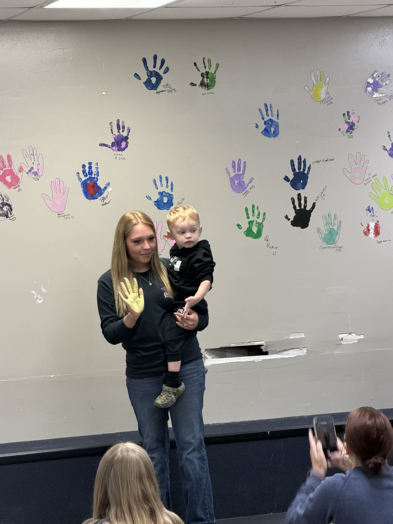 A woman holds a toddler on her hip, standing in front of a wall with many multicolored handprints.