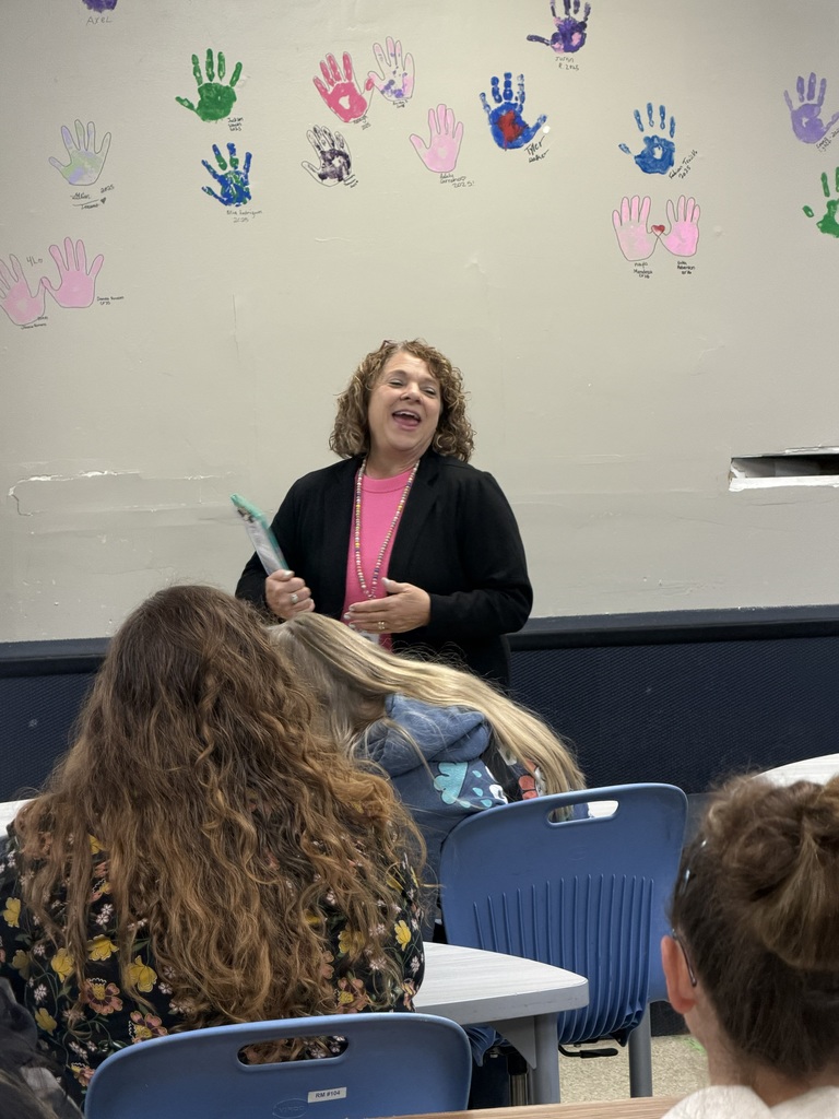 A woman in a pink shirt speaks in front of a classroom with handprints on the wall.