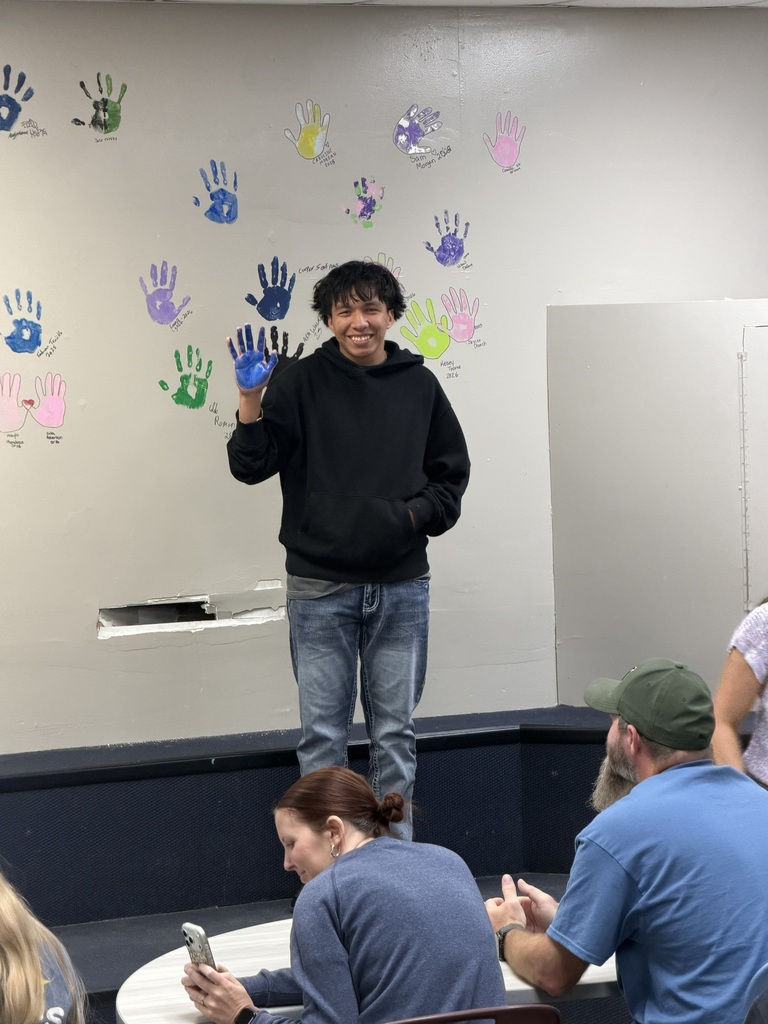 A man stands on a platform, wearing a black hoodie and jeans. He holds up blue-gloved hands. People are seated on chairs around him.