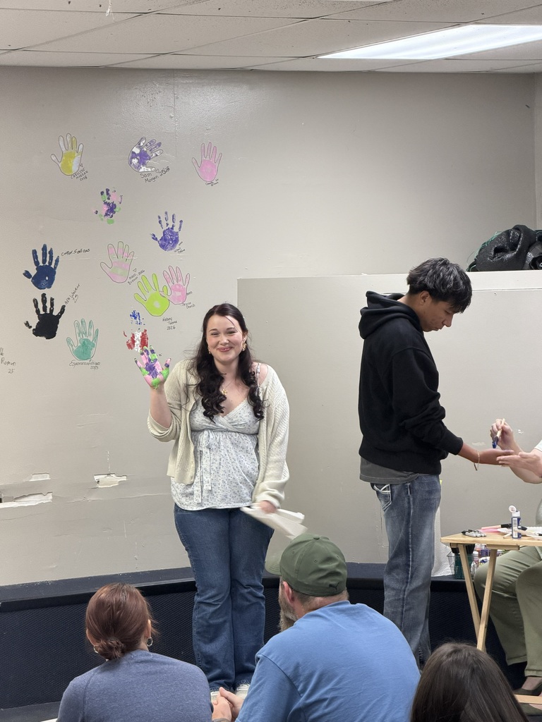 People stand and sit in a room. A woman raises her hand, and a man looks at her. A wall with painted hands is behind them.