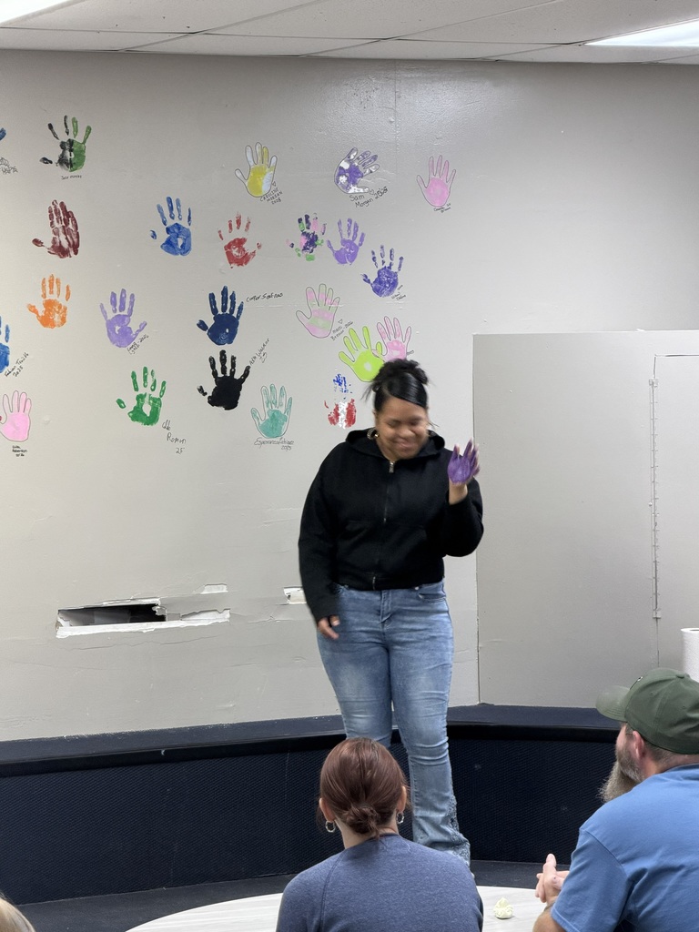 Woman in black jacket and jeans stands on a stage. Handprints in various colors decorate the wall behind her.
