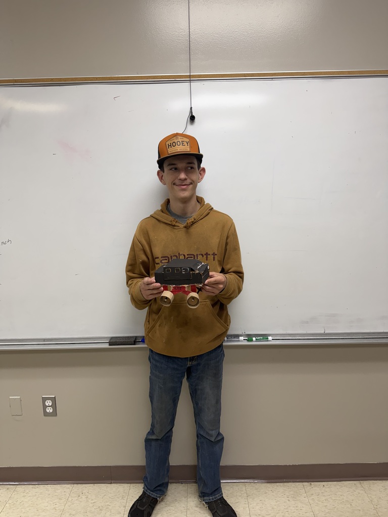 A person stands in a classroom, holding a small wheeled vehicle, wearing a hat and brown hoodie. A whiteboard is behind them.