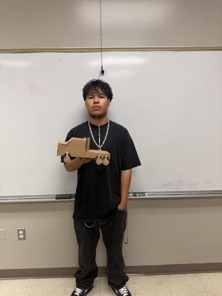 A man stands in a classroom, holding a wooden toy train in his hands. He wears a black shirt and pants.