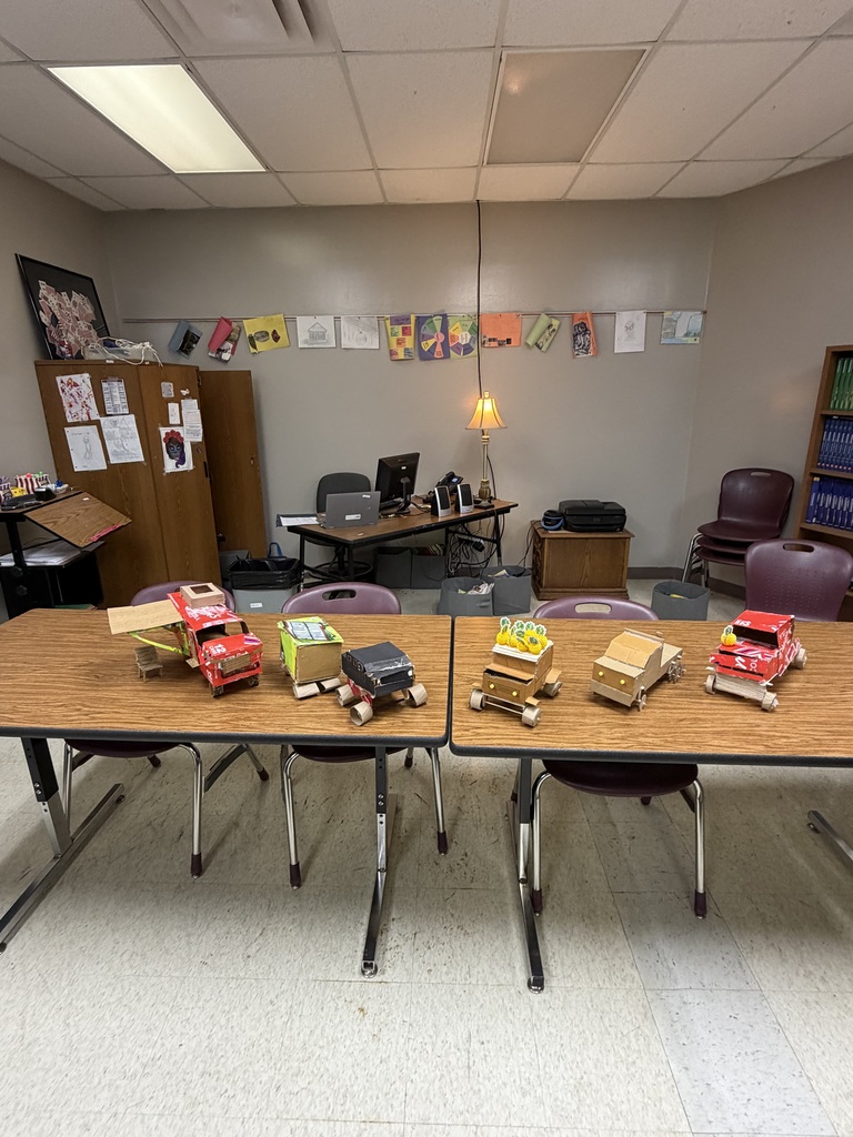 Classroom with tables and chairs. Toys on tables, a bookshelf, a lamp, and bulletin board. Desks and a computer are in the background.