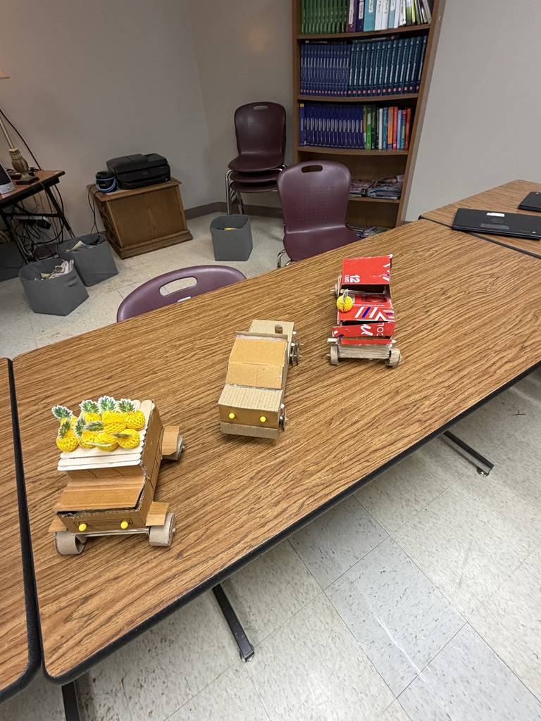 Classroom with desks and chairs. Three small toy trucks are on the table in the front.