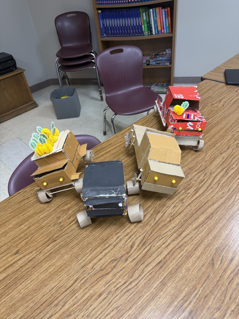 Four cardboard vehicles on a table in front of a bookshelf. A trash can is nearby.
