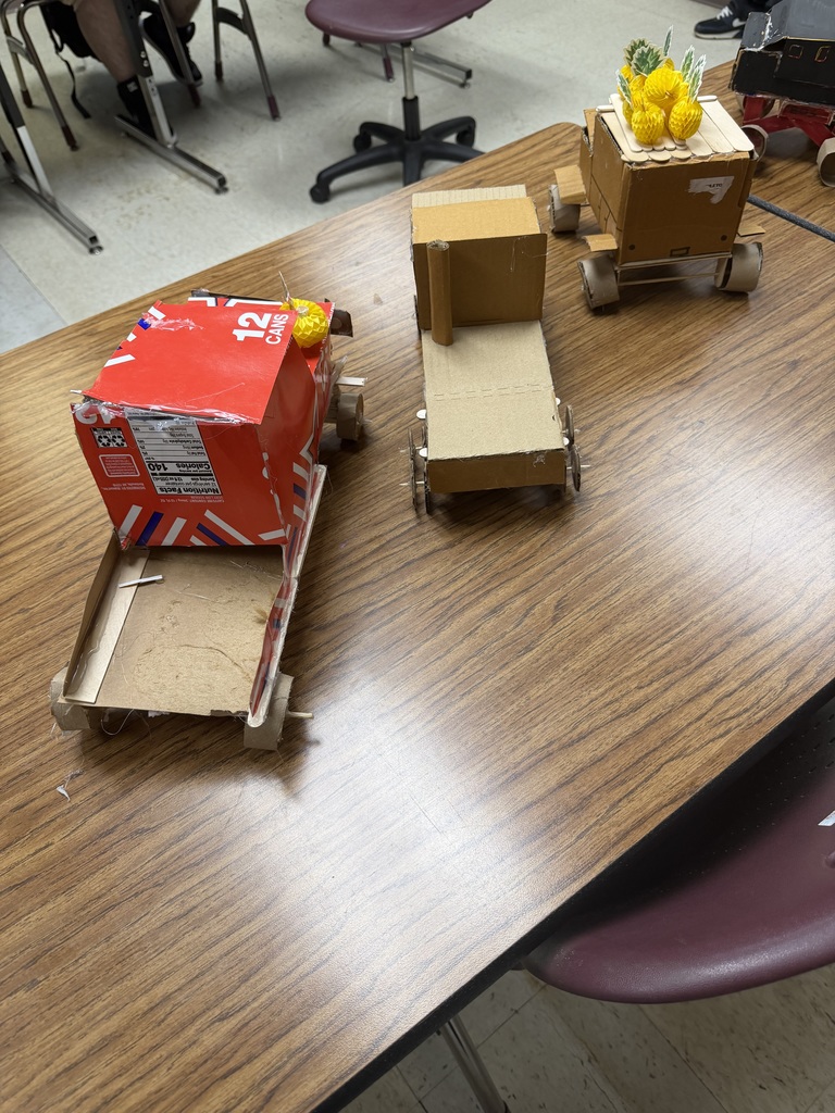 Four cardboard vehicles on a table. One has a red box on it. Chairs and a person's legs are visible.