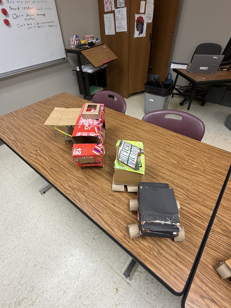 A classroom with a wooden table, chairs, and a whiteboard. On the table, there are several cardboard boxes and a black object.