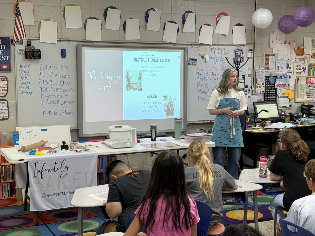 A classroom with a projector screen, a teacher, and students. The teacher is wearing an apron and the students are seated.