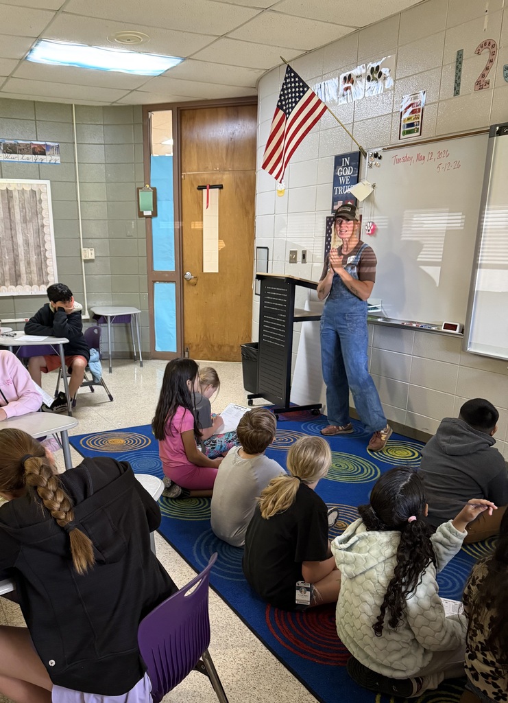 An indoor classroom with a famer standing, speaking to a group of seated students. A whiteboard is on the wall.