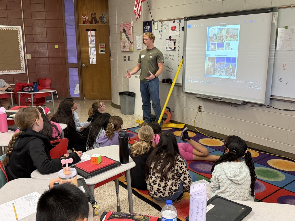 Man standing in front of a projector screen teaching kids in a classroom. Students sit on the floor.