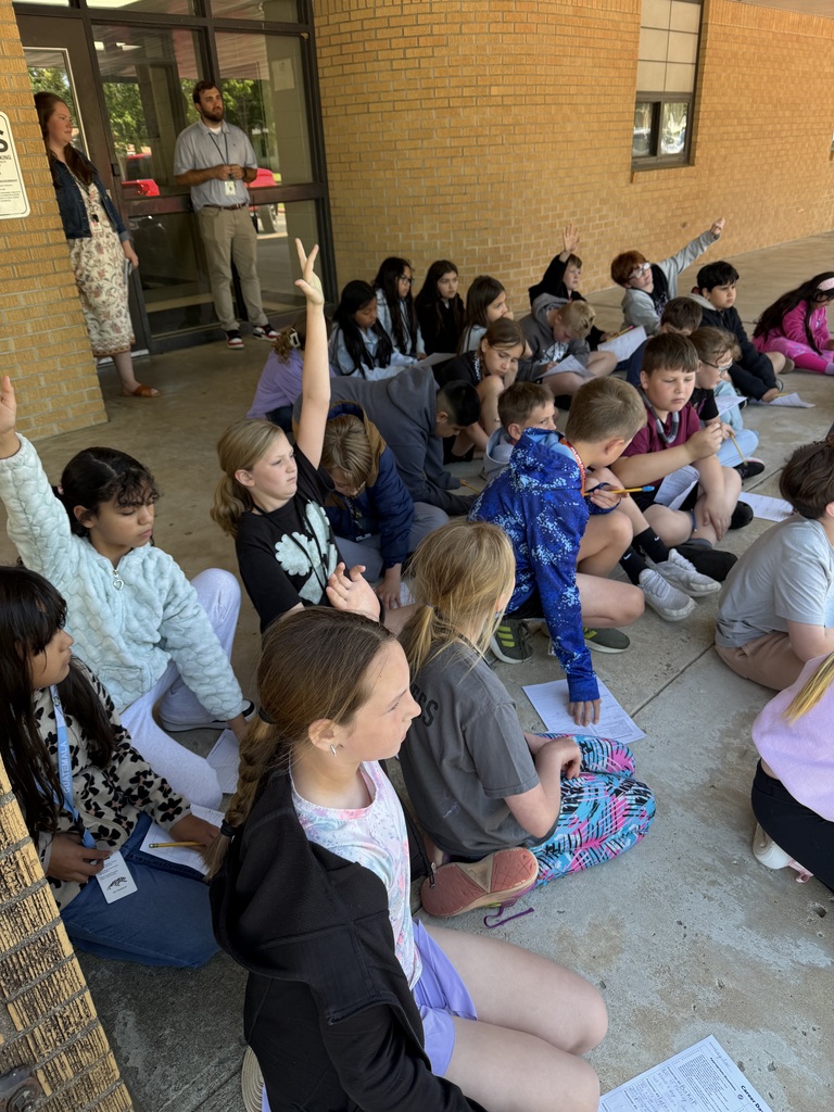A group of students sits on the floor, some raising hands, in front of a brick building.