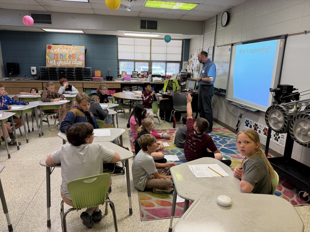 A classroom with students sitting at desks, some on rugs, facing a teacher presenting at the front with a projector screen.