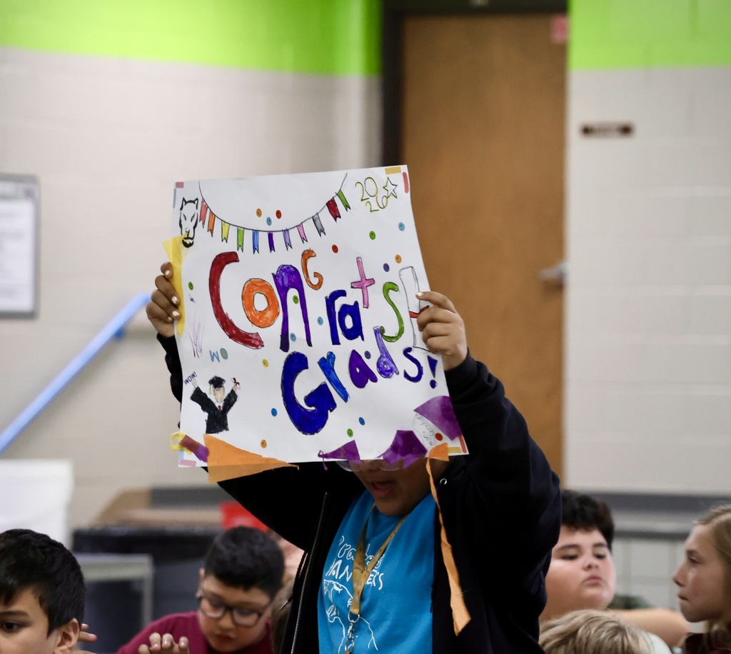 A person holding a colorful sign with the words "Congrats Grads!" inside a room with green walls.