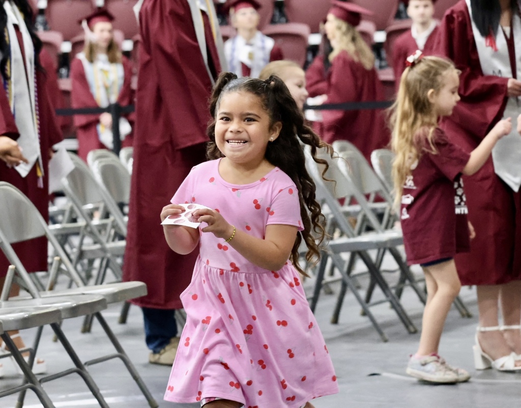 A young girl in a pink dress smiles, holding a certificate, surrounded by people in maroon gowns.
