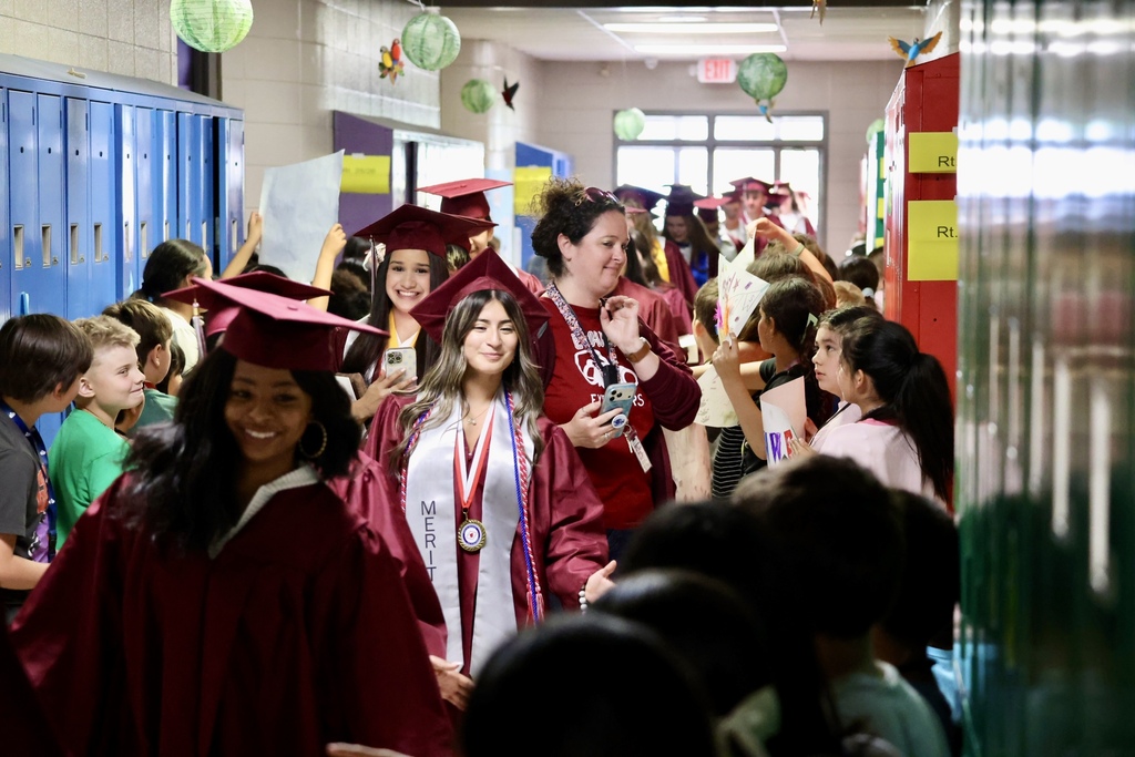 A school hallway filled with children in graduation gowns and caps, some walking and others watching.