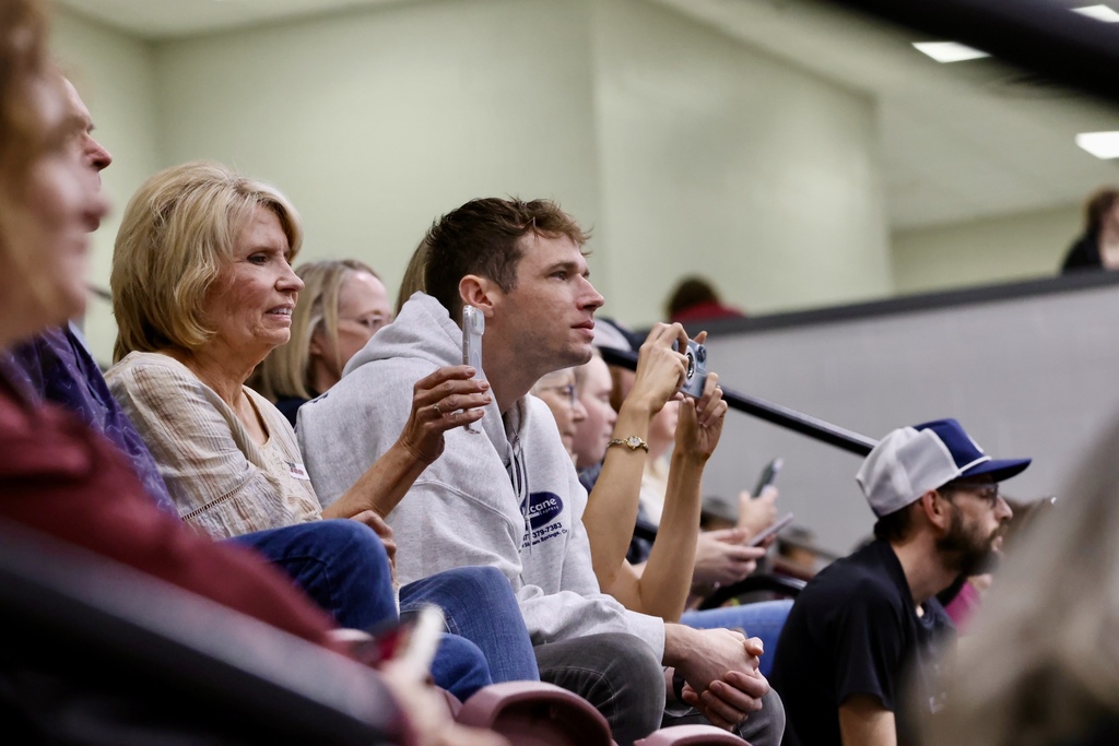 Several people in a stadium watching a presentation. Some are using their cell phones.