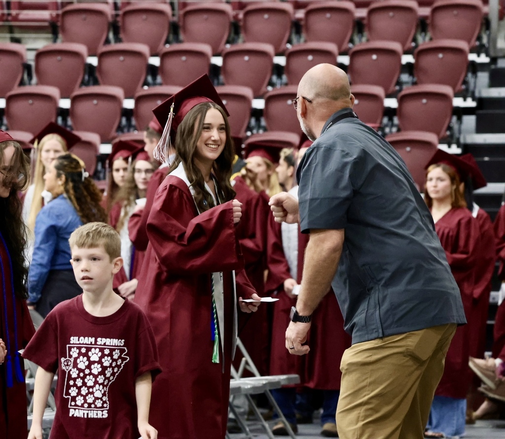 A person hands a diploma to a graduate in a maroon gown. A young boy and an adult look on.