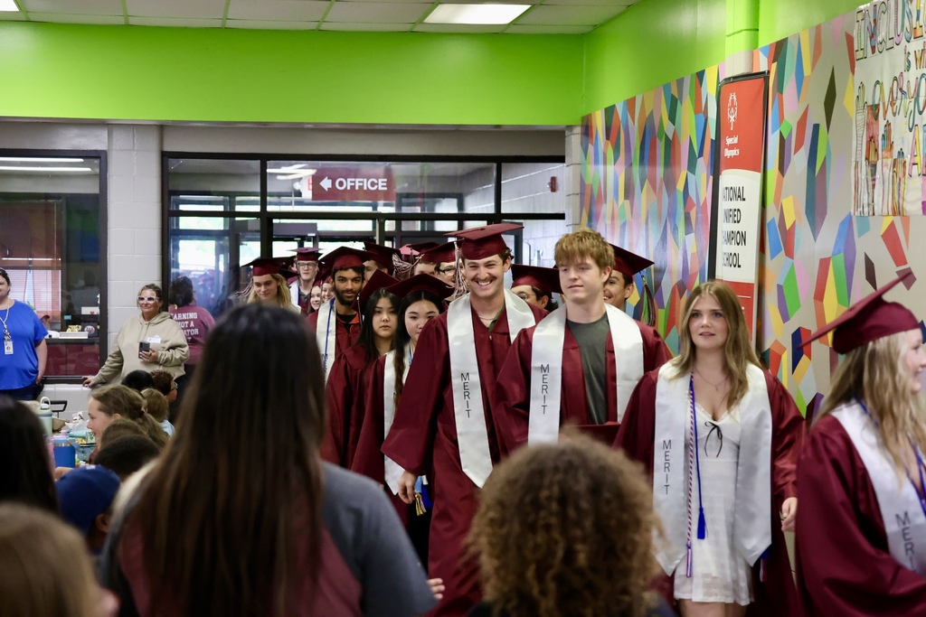 A line of graduates in caps and gowns walk through a school hallway, with colorful murals on the wall.