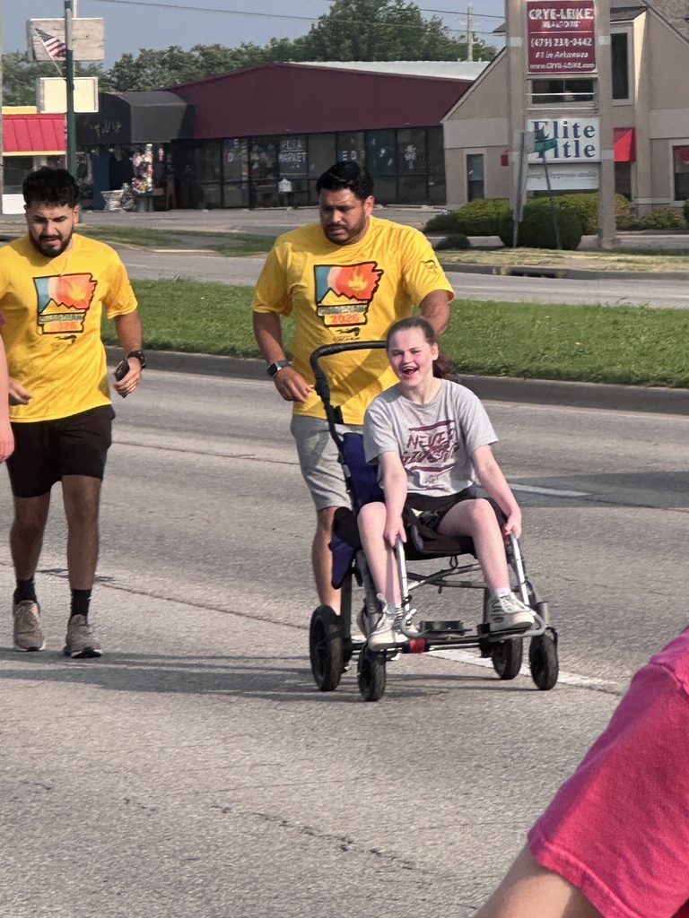 Two men in yellow shirts and a young girl in a wheelchair are walking on a street. Behind them, buildings and a grassy area.
