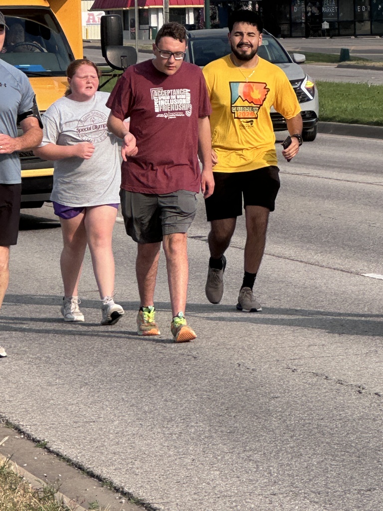 Four individuals walk in a street with a yellow bus and a parked car in the background. They wear casual clothing.