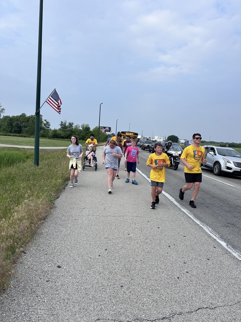A group of people running on a sidewalk, one pushing a wheelchair, with cars escorting them. An American flag is on a pole.