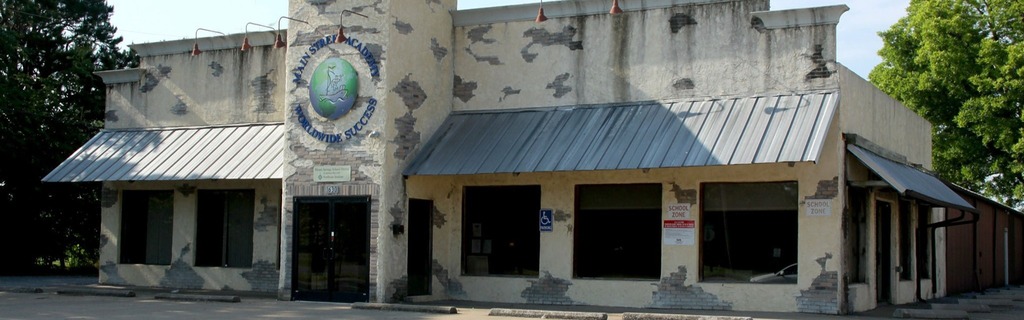 A white building with a metal roof, large windows, and a circular emblem on the front. Trees are in the background.