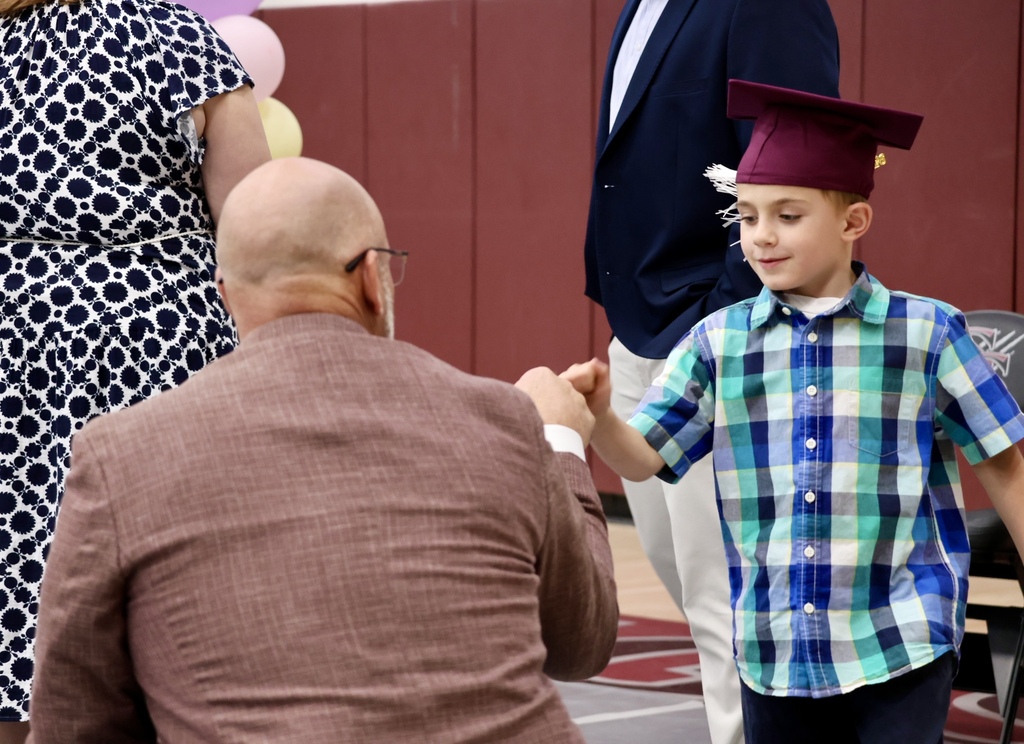 A man in glasses kneels, holding the hand of a young boy wearing a cap and plaid shirt.