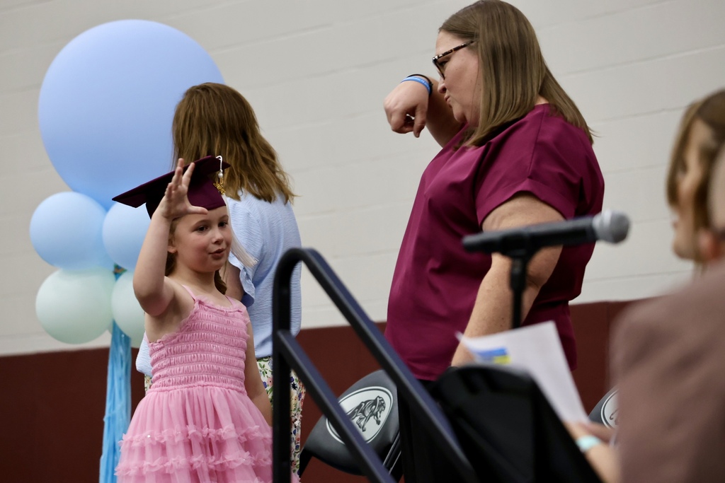 A young girl in a pink dress wears a graduation cap. A woman stands nearby.