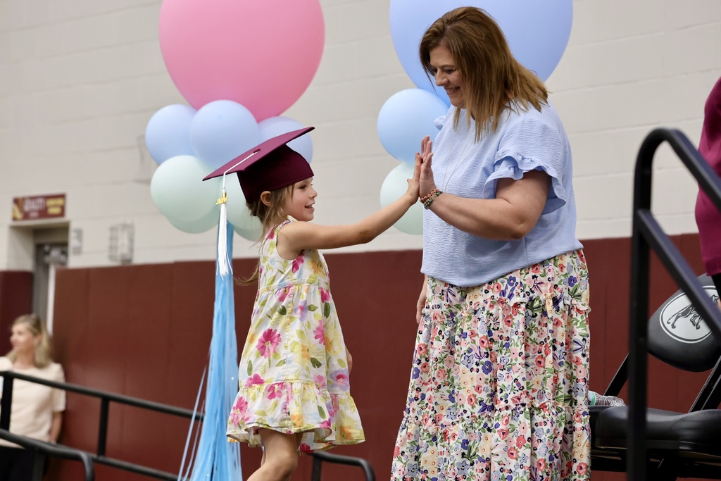 A woman and a young girl, both smiling, stand together in a room with balloons. The girl wears a graduation cap.