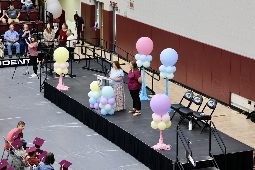 A stage with two women stands at a podium with balloons. Audience members sit in the bleachers.