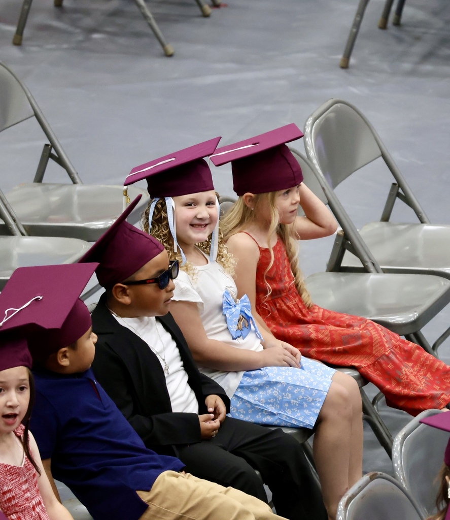 A group of children wearing graduation hats and gowns sit in chairs at a school event.